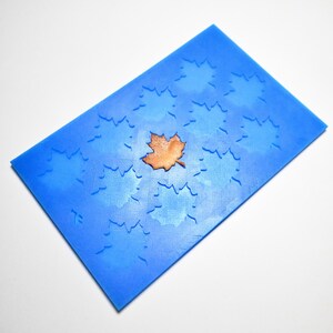 Puede incluir: Un molde de silicona azul con un diseño de hojas. El molde tiene una superficie texturizada con huellas de hojas y una sola hoja marrón en el centro. El molde es rectangular e inclinado, sobre un fondo blanco.
