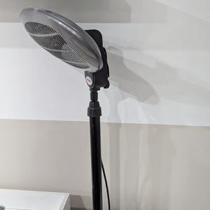 Op de afbeelding: Een zwarte en grijze verstelbare standventilator met een zilveren ventilatorkop. De ventilator is gemonteerd op een zwarte paal en staat op een wit oppervlak. De ventilatorkop heeft een roosterontwerp.
