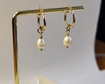 Puede incluir: Pendientes de aro dorados con una perla blanca colgante. Los pendientes se exhiben en un soporte de joyería dorado sobre un fondo neutro. Las perlas tienen un aspecto liso y brillante.