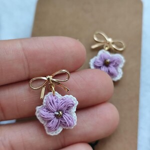 Könnte beinhalten: Ein Paar gehäkelte Blumenohrringe in Lila und Weiß mit goldenen Schleifenakzenten.