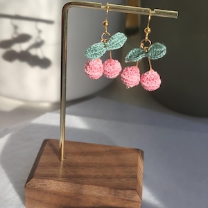 May include: A pair of crocheted cherry earrings with green leaves hanging from a gold stand. The cherries are pink and the leaves are a light green.