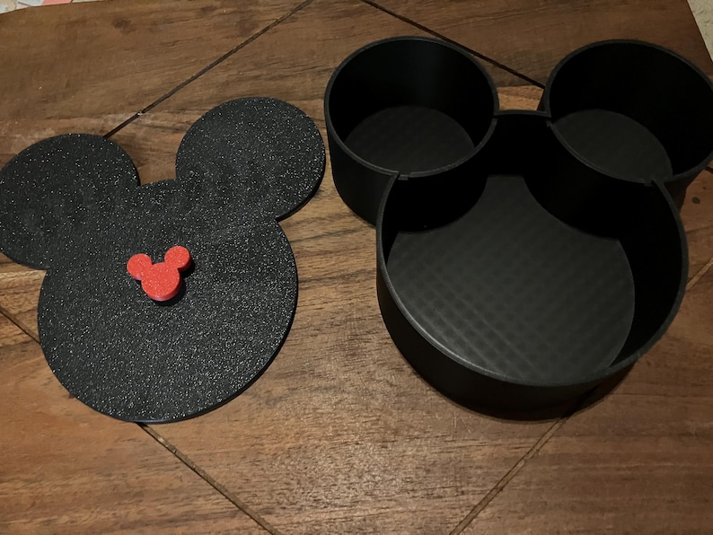 K&ouml;nnte beinhalten: Schwarzer Mickey-Maus-f&ouml;rmiger Aufbewahrungsbeh&auml;lter mit drei F&auml;chern. Der Deckel ist ein separates Teil mit einem roten Mickey-Maus-Kopf in der Mitte.