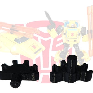 May include: Two black plastic pieces with a wavy design and a round center post. The pieces are designed to hold a toy robot's weapon.