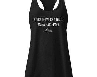 Puede incluir: Camiseta sin mangas negra con el texto blanco "STUCK BETWEEN A WALK AND A HARD PACE" y un gráfico de zapatilla blanca. La camiseta sin mangas tiene cuello redondo y está hecha de un material suave.