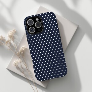 May include: A navy blue phone case with a white polka dot pattern. The case has a camera cutout and is resting on a white surface. A sprig of dried flowers is in the foreground.