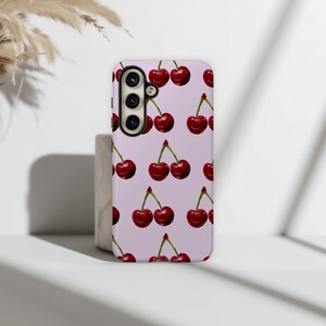 May include: A phone case with a repeating pattern of red cherries on a light purple background. The case has a camera module with three lenses and a flash. The cherries have green stems and are arranged in pairs.