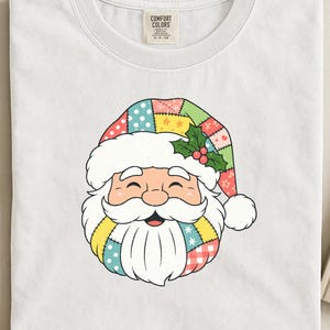 Op de afbeelding: Wit T-shirt met een kleurrijk ontwerp van een kerstman. Het gezicht van de kerstman heeft een patchwork hoed met stippen en hulst, een donzige witte baard en een glimlachende uitdrukking. Het shirt is gemaakt van een zacht materiaal.