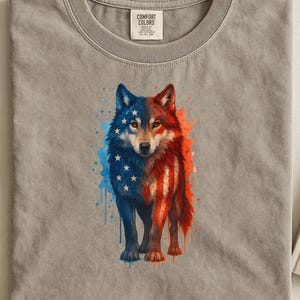 May include: A gray t-shirt featuring a wolf design with a patriotic theme. The wolf is split, with one side in blue with white stars and the other in red and white stripes, resembling the American flag. The shirt has a crew neck.