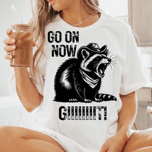 May include: White t-shirt featuring a black and white illustration of a raccoon wearing a cowboy hat and roaring. The text "GO ON NOW GUUUUUIT!" is printed above and below the raccoon graphic. The shirt is made of a soft material.