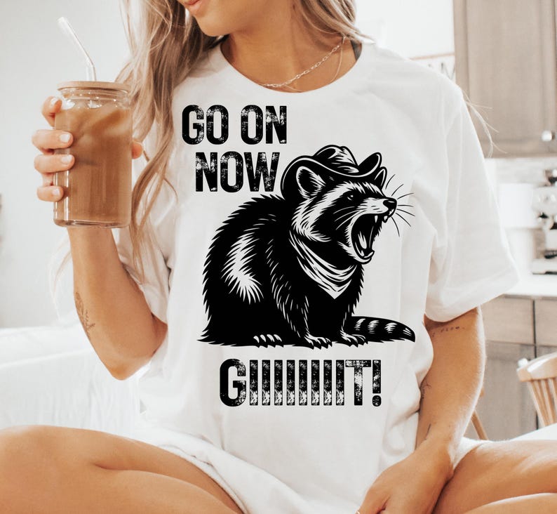 May include: White t-shirt featuring a black and white illustration of a raccoon wearing a cowboy hat and roaring. The text "GO ON NOW GUUUUUIT!" is printed above and below the raccoon graphic. The shirt is made of a soft material.