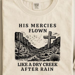 Op de afbeelding: Crèmekleurig T-shirt met een grafisch ontwerp met een kruis, cactussen en een woestijnlandschap. De tekst luidt "HIS MERCIES FLOWN LIKE A DRY CREEK AFTER RAIN". Het shirt heeft een Comfort Colors-label.
