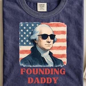 Op de afbeelding: Marineblauw T-shirt met een illustratie van George Washington met zonnebril voor een Amerikaanse vlag. De tekst "FOUNDING DADDY" is rood gedrukt. Het shirt heeft een ronde hals en een Comfort Colors label.