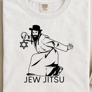 Op de afbeelding: Wit T-shirt met een zwarte afbeelding van een man in traditionele Joodse kleding, met een Davidster. De tekst "JEW JITSU" staat onder de illustratie. Het shirt heeft een ronde hals en een label met "Comfort Colors".