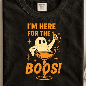 May include: Black t-shirt with a Halloween graphic. The design shows a cartoon ghost in a cocktail glass, holding a bottle. Orange text reads "I'm here for the BOOS!" The shirt has a Comfort Colors tag. A fun, spooky design.