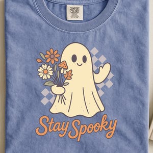 Op de afbeelding: Blauw Comfort Colors T-shirt met een crème cartoon spookje met een boeket bloemen en paddenstoelen. Het spookje heeft een lachend gezicht. De woorden "Stay Spooky" staan eronder in oranje gedrukt.
