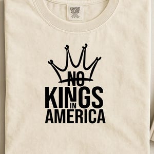 May include: A cream-coloured t-shirt featuring a black crown graphic and the text "NO KINGS IN AMERICA". The shirt has a crew neck and is made of a soft material. A casual tee for everyday wear.