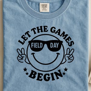 Op de afbeelding: Lichtblauw T-shirt met een zwarte afbeelding. De afbeelding toont een smiley met een zonnebril met "FIELD DAY" op de glazen. De tekst "LET THE GAMES BEGIN" staat rond het gezicht.