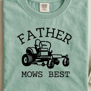 Op de afbeelding: Lichtgroen T-shirt met de zwarte tekst "FATHER MOWS BEST". Het ontwerp toont een zwarte illustratie van een zitmaaier. Het shirt is van het merk Comfort Colors.
