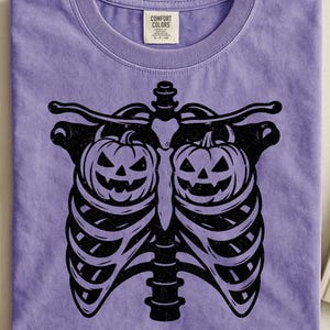 Op de afbeelding: Een lila Comfort Colors T-shirt met een zwarte afbeelding van een ribbenkast en twee pompoenen. Het T-shirt met ronde hals heeft een label op de kraag. Een Halloween-thema ontwerp.