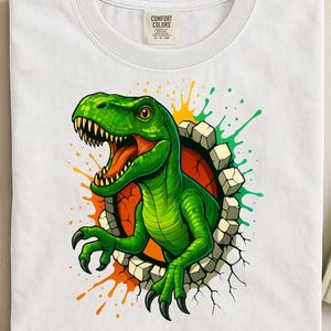 Op de afbeelding: Wit T-shirt met een levendige groene dinosaurus die door een muur breekt. De dinosaurus heeft scherpe tanden en klauwen, met oranje en groene verfspatten. Het shirt heeft een Comfort Colors label.