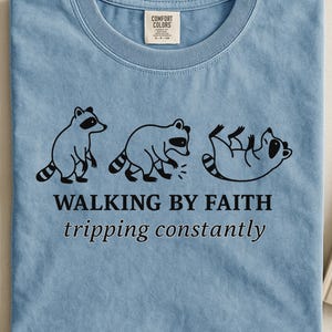 Op de afbeelding: Lichtblauw T-shirt met een afbeelding van drie wasberen in verschillende poses. De tekst "WALKING BY FAITH tripping constantly" staat eronder. Het T-shirt is gemaakt van zacht materiaal.