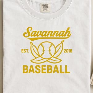 Op de afbeelding: Wit T-shirt met een gouden ontwerp met het woord "Savannah" boven een honkbalafbeelding en het woord "BASEBALL" eronder. Het ontwerp bevat "EST. 2016". Het shirt is opgevouwen.