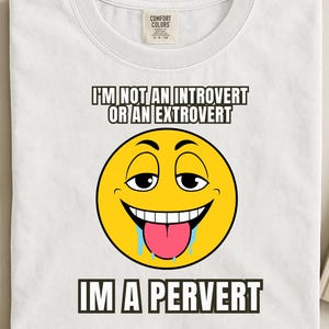 Op de afbeelding: Wit T-shirt met een gele emoji-gezichtsgrafiek. De emoji heeft een brede glimlach, een roze tong en kwijl. Boven de emoji staat de tekst "I'M NOT AN INTROVERT OR AN EXTROVERT" en daaronder "IM A PERVERT".