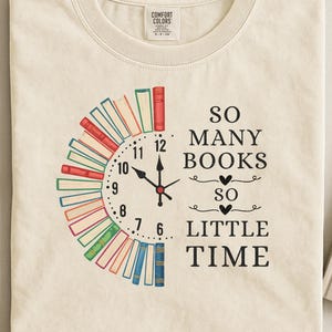 Op de afbeelding: Crèmekleurig T-shirt met een klokontwerp gemaakt van boeken. De wijzers van de klok wijzen naar 12:00 uur. De tekst luidt "So Many Books So Little Time". Een boek is rechts zichtbaar.