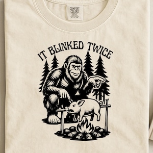 May include: Cream-colored t-shirt featuring a black graphic of a Bigfoot figure pointing at a pig roasting over a campfire. The text "IT BLINKED TWICE" is arched above the illustration. The shirt is made of a soft material.