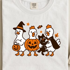 Op de afbeelding: Wit T-shirt met een Halloween-thema. Het ontwerp toont cartoonkippen in kostuums, een zwarte kat, een pompoen en snoep. Het shirt heeft een Comfort Colors label.