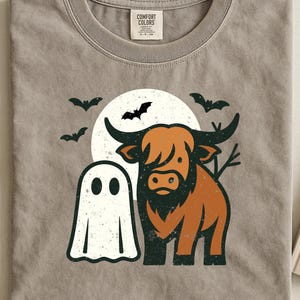 Op de afbeelding: Lichtbruin T-shirt met een Halloween-thema. Het ontwerp bevat een cartoongeest, een Schotse hooglander, vleermuizen en een volle maan. Het shirt heeft een ronde hals en een Comfort Colors-label.
