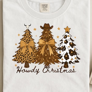Op de afbeelding: Wit T-shirt met drie kerstbomen met een western thema. De bomen zijn versierd met luipaardprint, cowboyhoeden en sterren. De tekst "Howdy Christmas" is geschreven in een sierlijk lettertype.