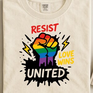 Op de afbeelding: Crèmekleurig T-shirt met een regenboogvuist-afbeelding. De woorden "RESIST" en "UNITED" staan in rood en wit, en "LOVE WINS" in geel. Het ontwerp bevat bliksemschichten en een zwarte inktspetter.