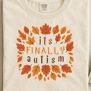 Op de afbeelding: Crèmekleurig T-shirt met ronde hals. De voorkant toont een cirkelvormig ontwerp van herfstbladeren in oranje en bruin, met de woorden "its FINALLY autism" in een sierlijk lettertype.
