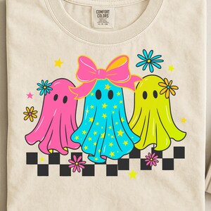 Op de afbeelding: Crèmekleurig T-shirt met een afbeelding van drie cartoongeesten. De geesten zijn roze, blauw en geel, versierd met strikken, bloemen en sterren. Onderaan staat een zwart-wit dambordpatroon.