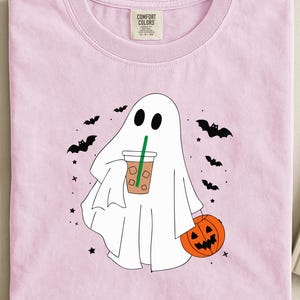 Op de afbeelding: Lichtroze T-shirt met een witte spookafbeelding met een koffiekopje en een pompoen. Zwarte vleermuizen en sterren omringen het spook. Het shirt heeft een ronde hals en een label met "COMFORT COLORS".