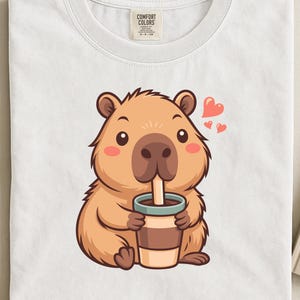 Op de afbeelding: Wit T-shirt met een cartoon capibara die uit een koffiekopje drinkt. De capibara is lichtbruin met een donkerbruine neus en roze hartjes. Het shirt heeft een Comfort Colors label.