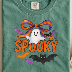Op de afbeelding: Een groen Comfort Colors T-shirt met een Halloween-ontwerp. Het ontwerp toont een witte geest, een oranje en paarse strik, oranje vleermuizen en het woord "SPOOKY" in oranje letters. Onderaan staat een zwarte vleermuis.