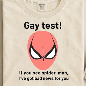 Puede incluir: Camiseta color crema con el texto "Gay test!" sobre un gráfico de máscara de Spider-Man rosa. Debajo del gráfico, el texto dice: "If you see spider-man, I've got bad news for you."
