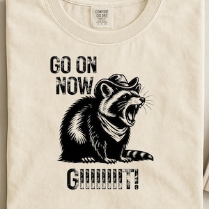 Op de afbeelding: Crèmekleurig T-shirt met een zwart-wit illustratie van een wasbeer met cowboyhoed en bandana, met de tekst "GO ON NOW GIIIIIIIT!" erboven en eronder. Het shirt heeft een ronde hals.