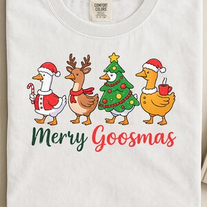 Op de afbeelding: Wit T-shirt met een kerstthema. Het toont vier cartoon ganzen in feestelijke kleding: een gans met een zuurstok, een rendiergans, een kerstboomgans en een gans met een mok. De tekst "Merry Goosmas" staat eronder.