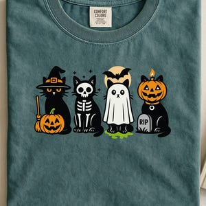 Op de afbeelding: Teal Comfort Colors T-shirt met een Halloween-thema grafiek. Het ontwerp bevat vier zwarte katten verkleed als heks, skelet, spook en pompoen. Een volle maan en een RIP grafsteen zijn ook inbegrepen.