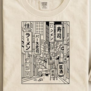 May include: Cream-colored t-shirt featuring a black and white illustration of a Japanese street scene. The design includes Japanese text on signs, buildings, a car, and a lantern, creating a detailed urban landscape.