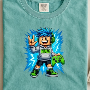 Op de afbeelding: Een turquoise Comfort Colors T-shirt met een cartoon game-personage. Het personage heeft blauw haar, een koptelefoon en een groene controller. Het personage draagt een hoodie met het woord "NINJA" en is omgeven door een blauw bliksemontwerp.