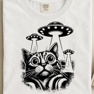 Könnte beinhalten: Weißes T-Shirt mit einer schwarz-weißen Grafik einer Katze, die von drei UFOs entführt wird. Die Katze hat große Augen und einen überraschten Gesichtsausdruck. Das Shirt hat ein Comfort Colors-Etikett.