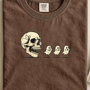 Puede incluir: Camiseta marrón con un gráfico de una calavera y tres pequeños fantasmas. La calavera es blanquecina con grietas, y los fantasmas son blancos con ojos negros. La camiseta tiene cuello redondo y una etiqueta en el centro superior.