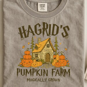 Op de afbeelding: Een grijs Comfort Colors T-shirt met een afbeelding van Hagrid's Pumpkin Farm. Het ontwerp bevat een huis, pompoenen en de tekst "Hagrid's Pumpkin Farm Magically Grown". Het shirt heeft een ronde hals en korte mouwen.