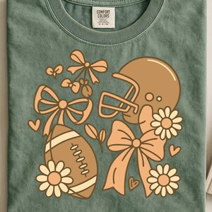 Op de afbeelding: Groen Comfort Colors T-shirt met een voetbal, helm, strikken en madeliefjes in bruin- en perziktinten. Het ontwerp heeft een vintage, retro esthetiek. Het shirt heeft een ronde hals en korte mouwen.