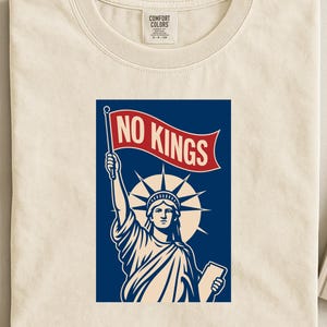 Op de afbeelding: Crèmekleurig T-shirt met een afbeelding van het Vrijheidsbeeld met een vlag met de tekst "NO KINGS" in rode letters. Het ontwerp is geplaatst tegen een blauwe achtergrond met een zonnestraaleffect.