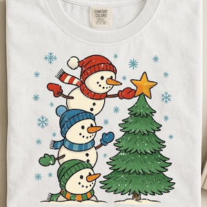 Op de afbeelding: Wit T-shirt met een illustratie van drie sneeuwmannen die een kerstboom versieren. De sneeuwmannen dragen rode, blauwe en groene hoeden en sjaals. Een gouden ster staat bovenop de groene boom, met sneeuwvlokken.
